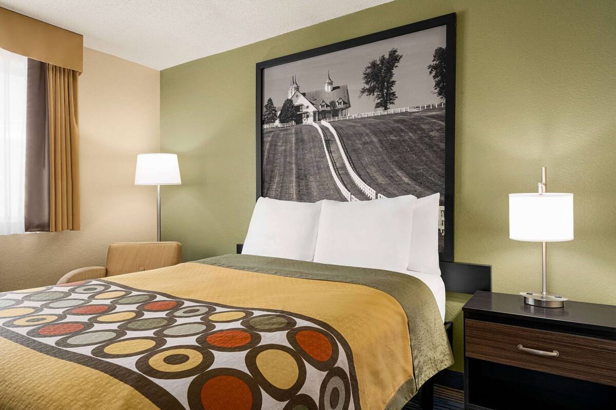 Habitación de hotel con cama, dos lámparas de noche, un cuadro grande en la pared y una mesita de noche con cajón.