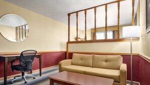 A hotel room with a sofa, a desk with a chair, a round mirror, a wooden ceiling railing, and a floor lamp.