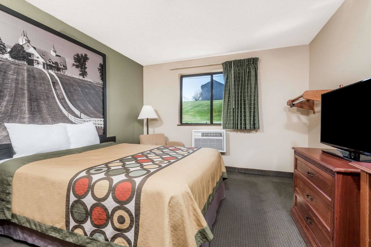 Habitación de hotel con cama, televisión, cómoda, ventana con cortinas, aire acondicionado y un cuadro en la pared.