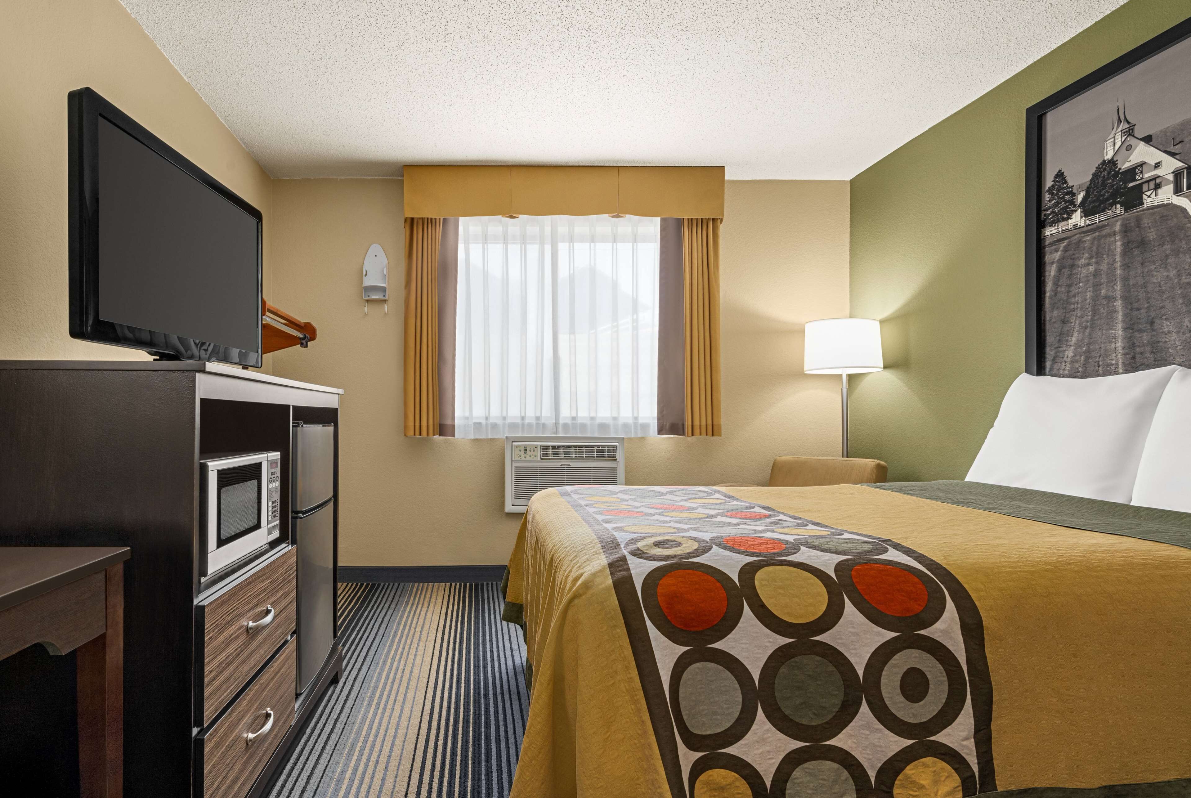 A hotel room with a bed, a television, a microwave, a dresser, a window with curtains, and a painting on the wall.