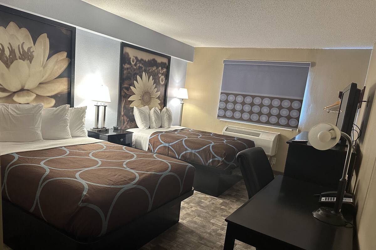 A hotel room with two beds, a desk, a chair, a lamp, and a large flower painting on the wall.
