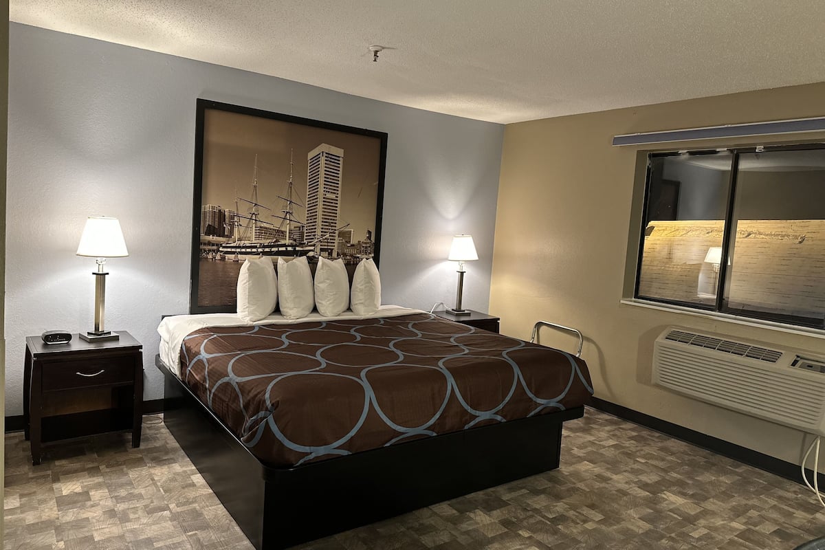 A hotel room with a bed, two bedside tables with lamps, a window with a view of the city, and a wall-mounted air conditioner.