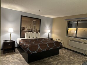 A hotel room with a bed, two bedside tables with lamps, a window with a view of the city, and a wall-mounted air conditioner.