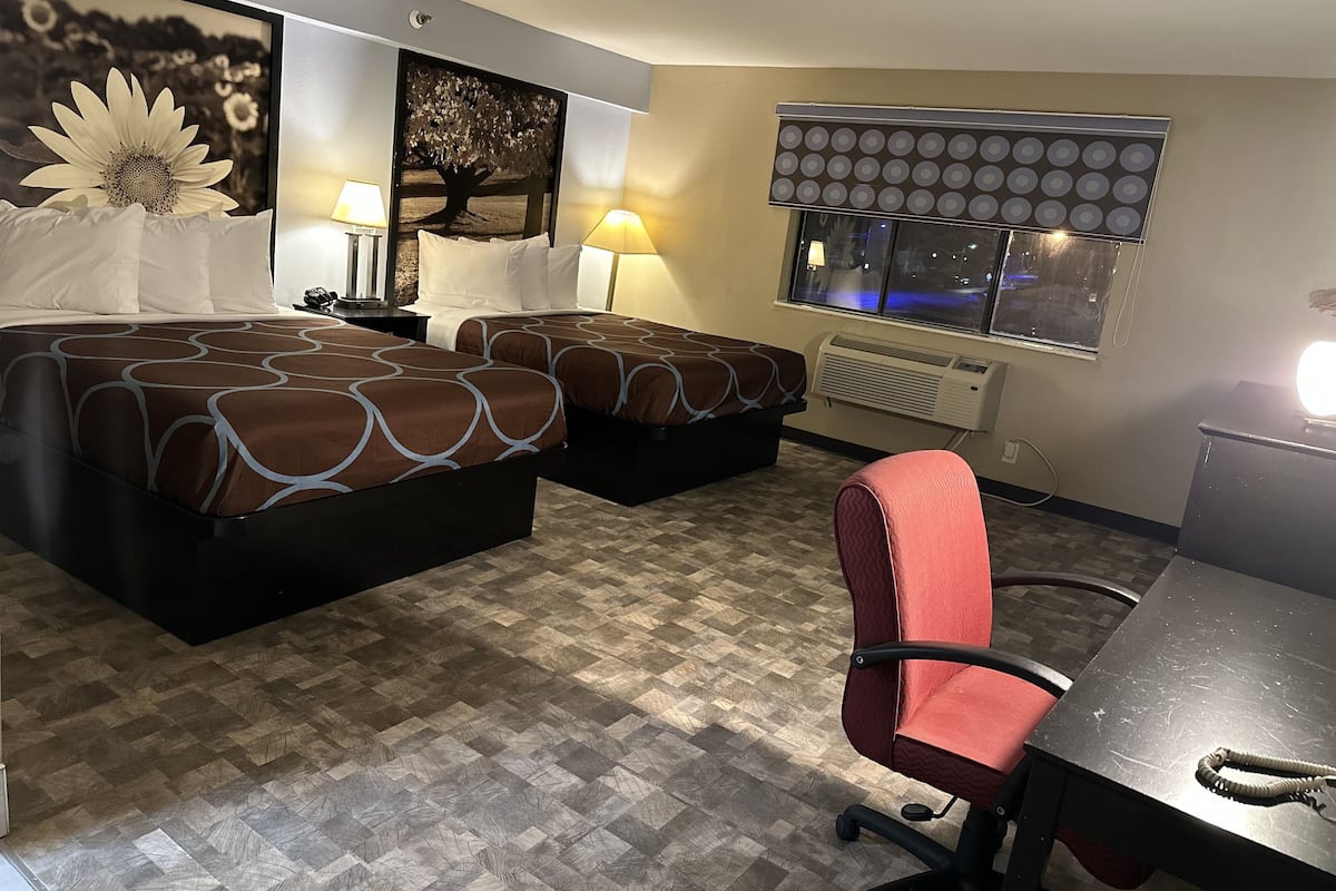 A hotel room with two beds, a desk, a chair, a window with blinds, and a wall-mounted air conditioner.