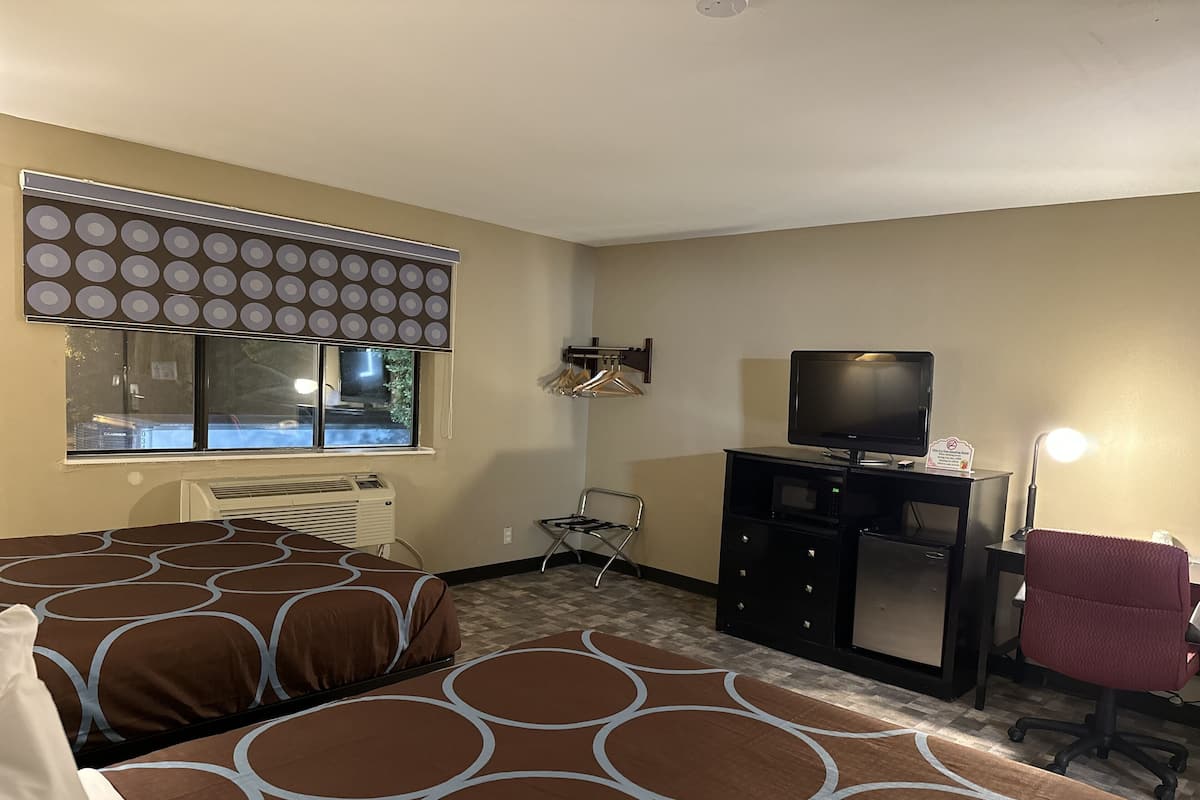 A hotel room with two beds, a desk with a television, a chair, a desk lamp, and a window with a view of the outside.