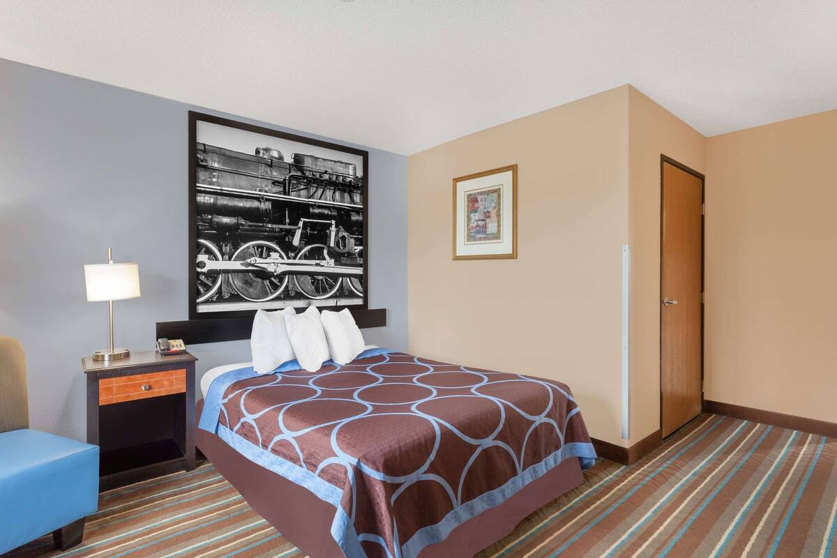 Quarto de hotel com cama, mesa de cabeceira, luminária, cadeira, quadro na parede e porta.