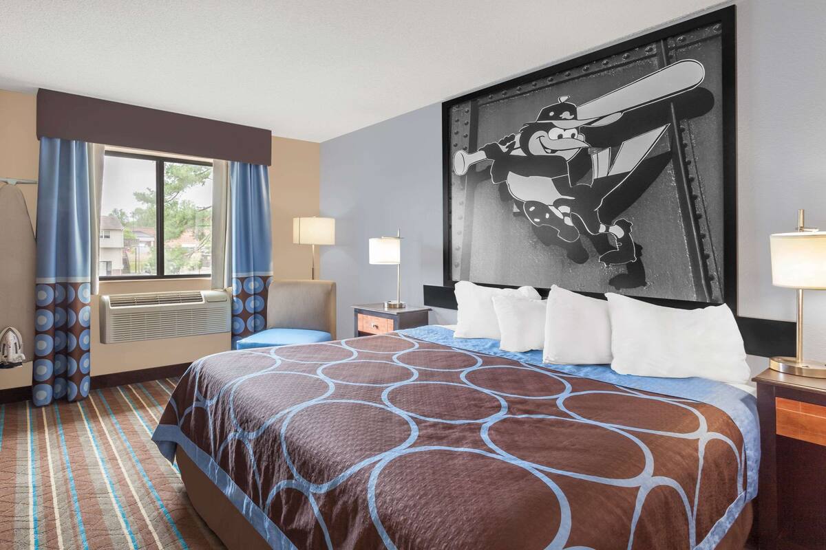 Quarto de hotel com cama grande, janela com cortinas, obra de arte na parede, mesa de cabeceira com luminária e cabeceira.