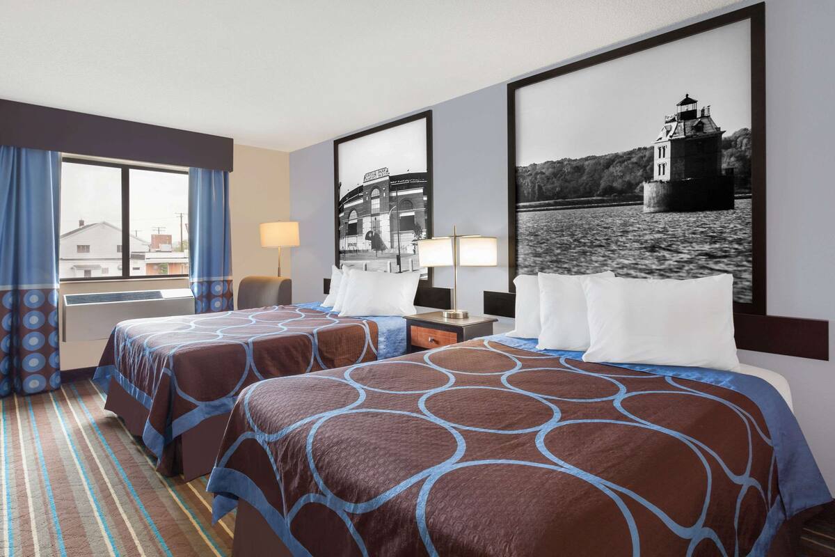 Quarto de hotel com duas camas, uma janela grande com cortinas, um quadro com uma imagem de um farol e uma luminária na mesa de cabeceira.