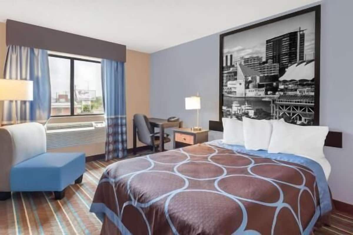 Quarto de hotel com cama, escrivaninha, cadeira, janela com cortinas e pintura da paisagem urbana na parede.