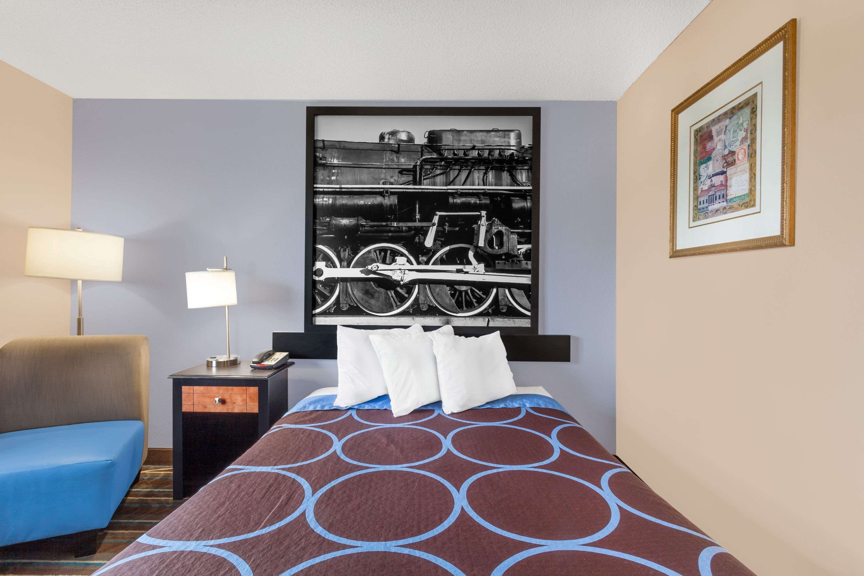 A hotel room with a bed, a chair, a nightstand with a lamp, and a framed picture on the wall.