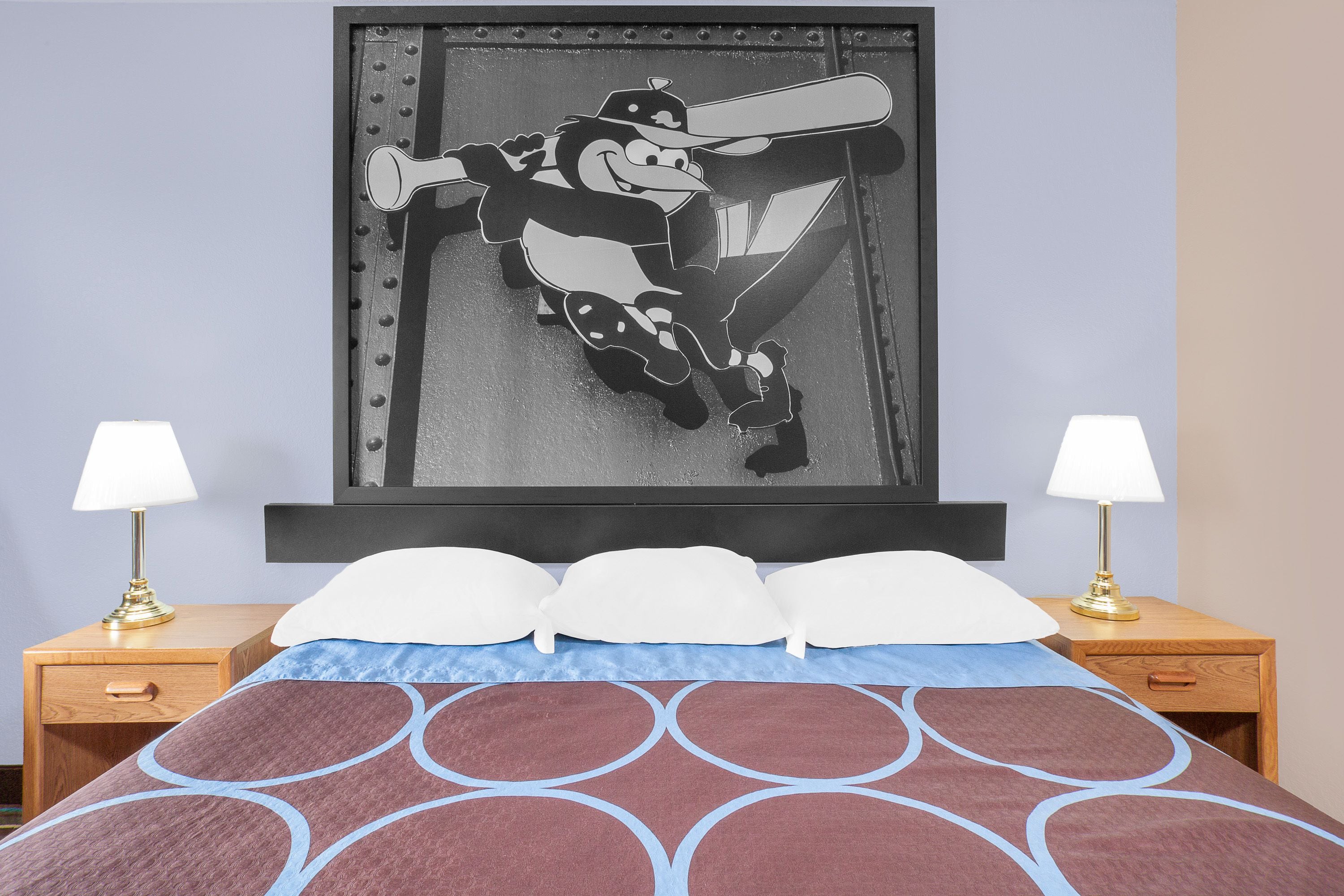 A bed with a patterned bedspread, two bedside lamps, a framed picture on the wall, and a wooden nightstand with a drawer.