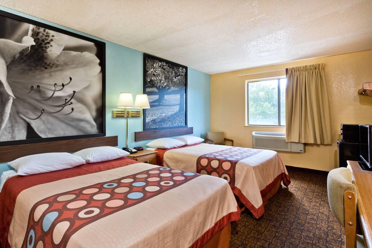 A hotel room with two beds, a large wall art of a flower, a window with curtains, and a television.