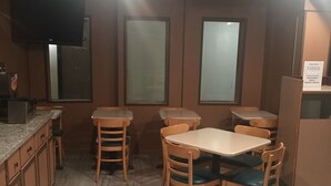 Un comedor con mesas y sillas de madera, una zona de cocina con armarios y un refrigerador, y un televisor fijado a la pared.