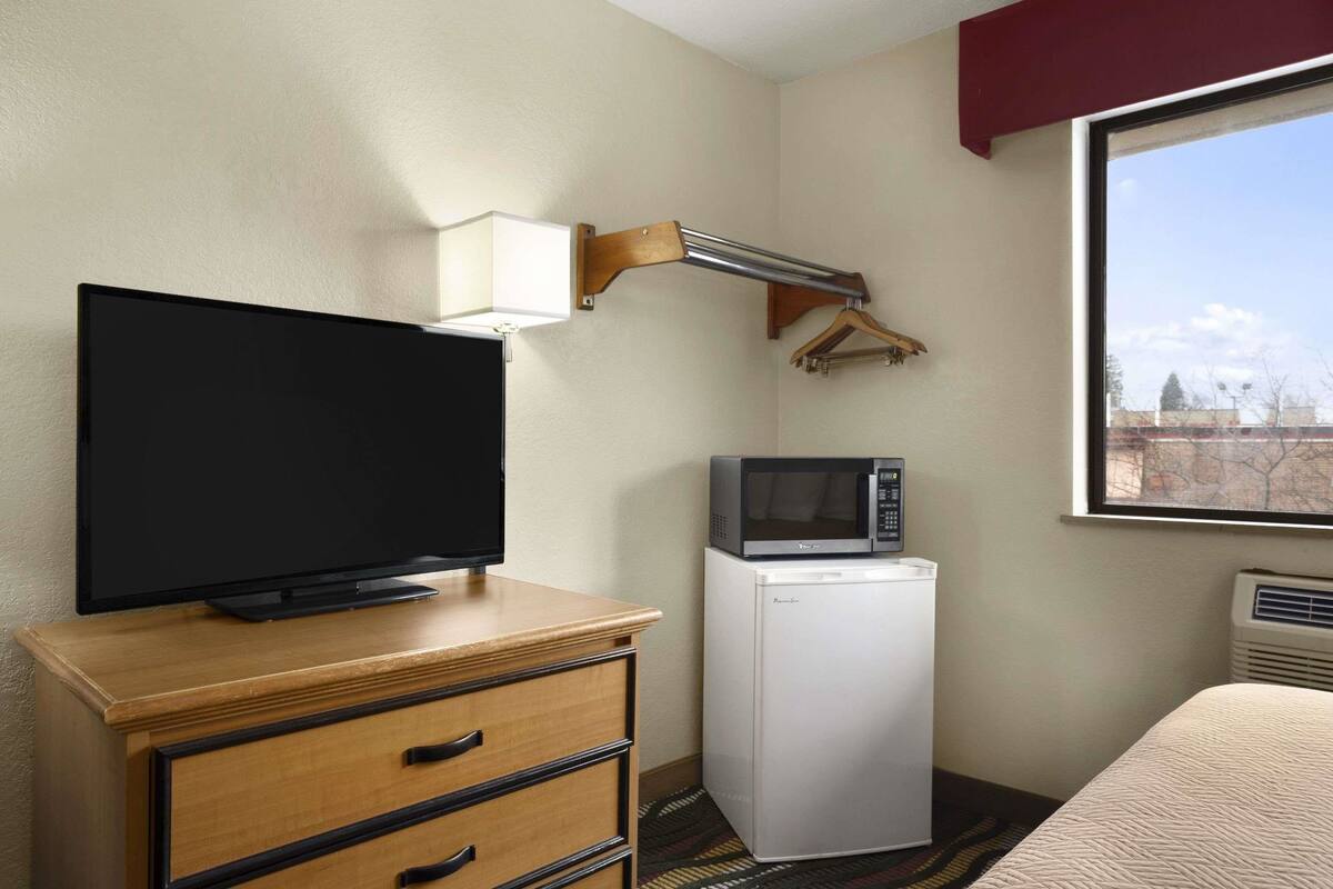 A hotel room with a television on a wooden dresser, a microwave, and a window with a view of buildings.