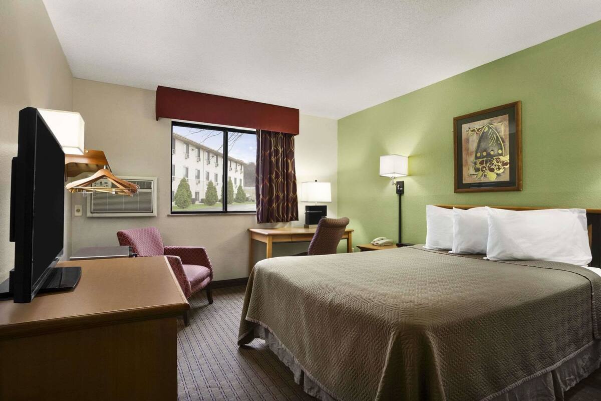 A hotel room with a bed, desk, chair, TV, window, lamp, and artwork.