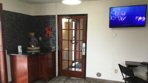 Una habitación con cocina, puerta de madera, televisor montado en la pared y una pequeña mesa con una silla.