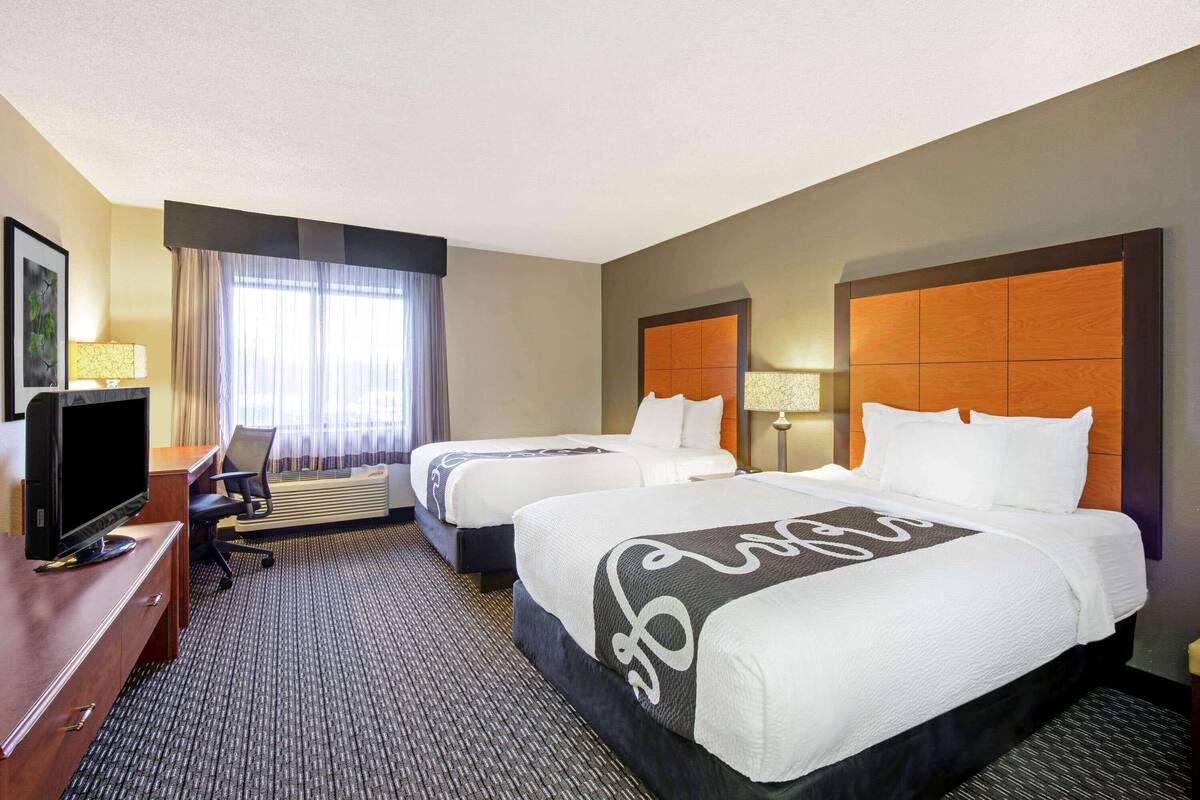 Ein Hotelzimmer mit zwei Betten, einem Schreibtisch mit Fernseher und einem großen Fenster mit Vorhängen.