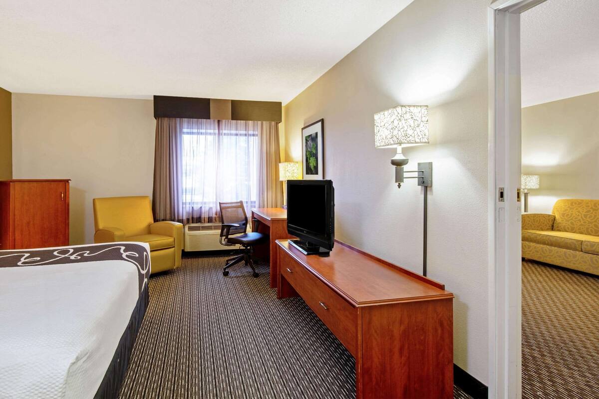 Ein Hotelzimmer mit Bett, Schreibtisch, Fernseher, Stuhl, Sofa, Lampe und einem Fenster mit Vorhängen.