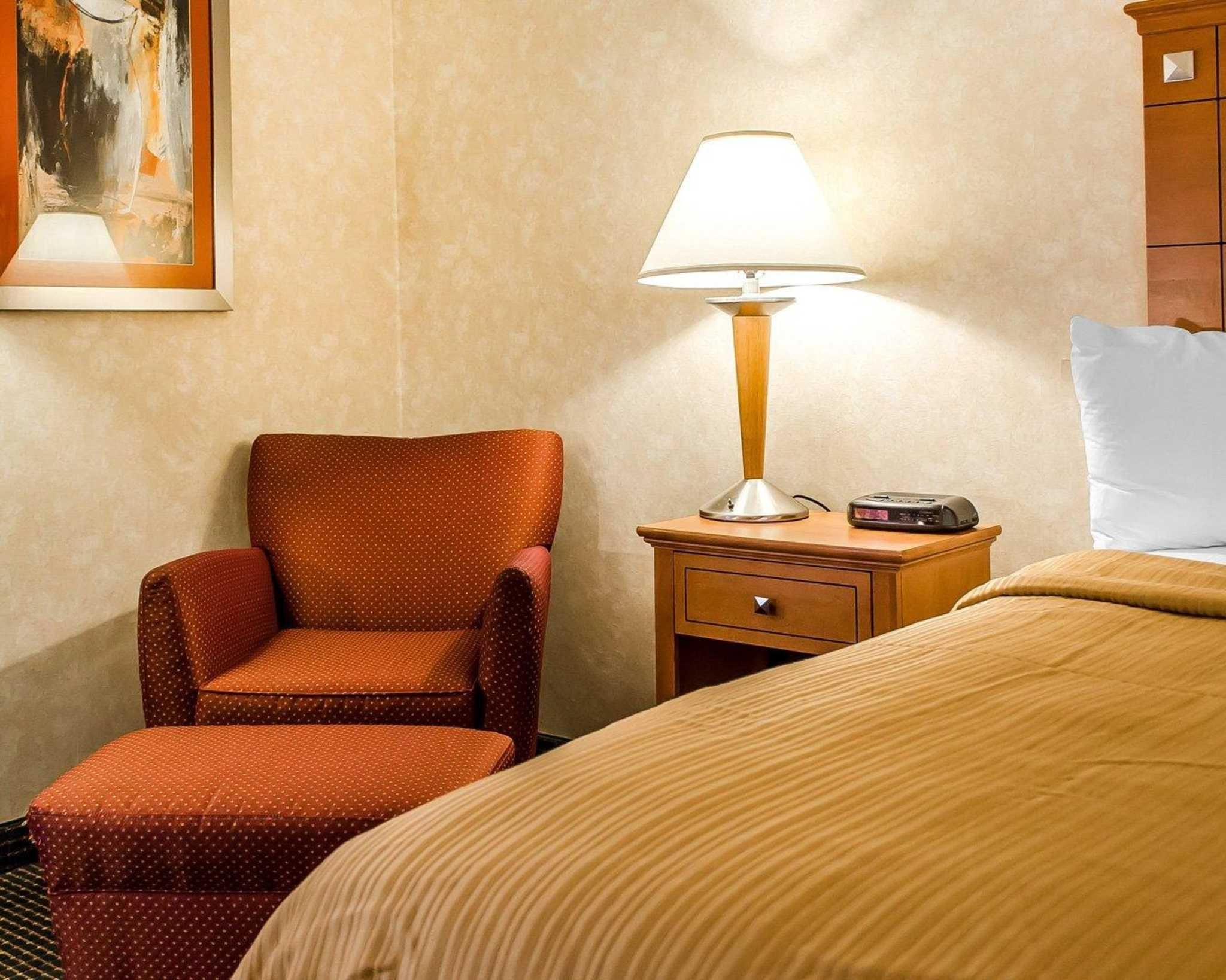 A hotel room with a bed, a chair, a nightstand with a lamp, and a painting on the wall.