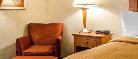 A hotel room with a bed, a chair, a nightstand with a lamp, and a painting on the wall.
