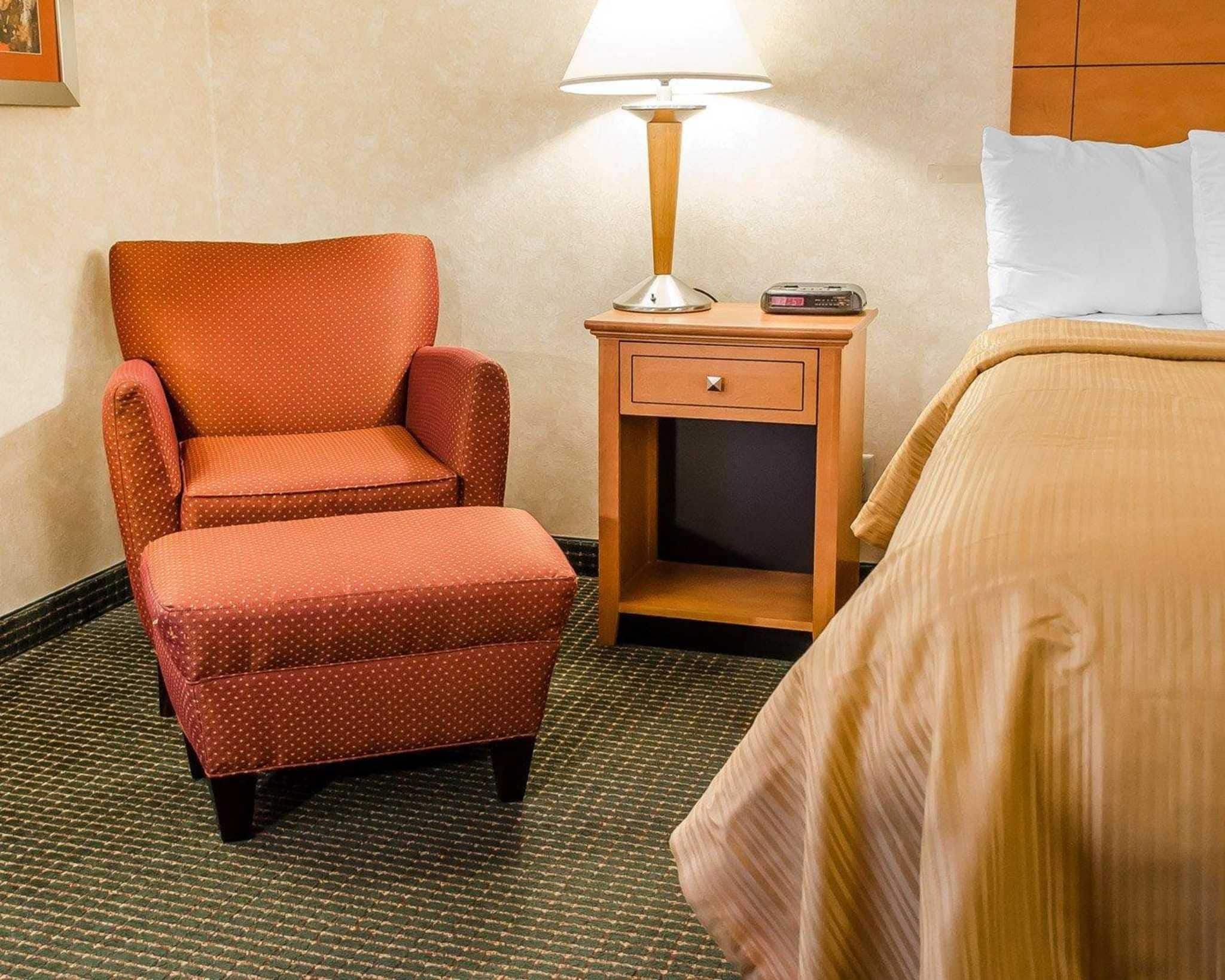 Una habitación de hotel con un sillón estampado de lunares, una mesita de noche con una lámpara, un reloj digital y una cama con almohadas blancas.