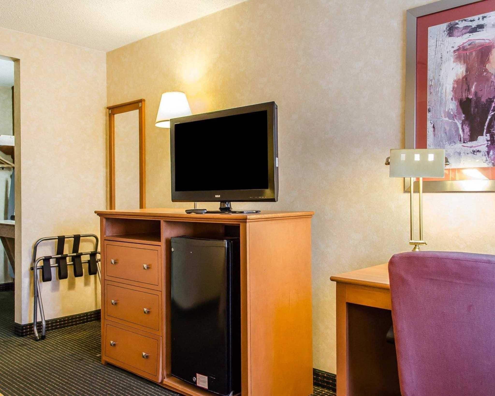 Habitación de hotel con un escritorio de madera, televisión, refrigerador, lámpara y cama con manta morada.