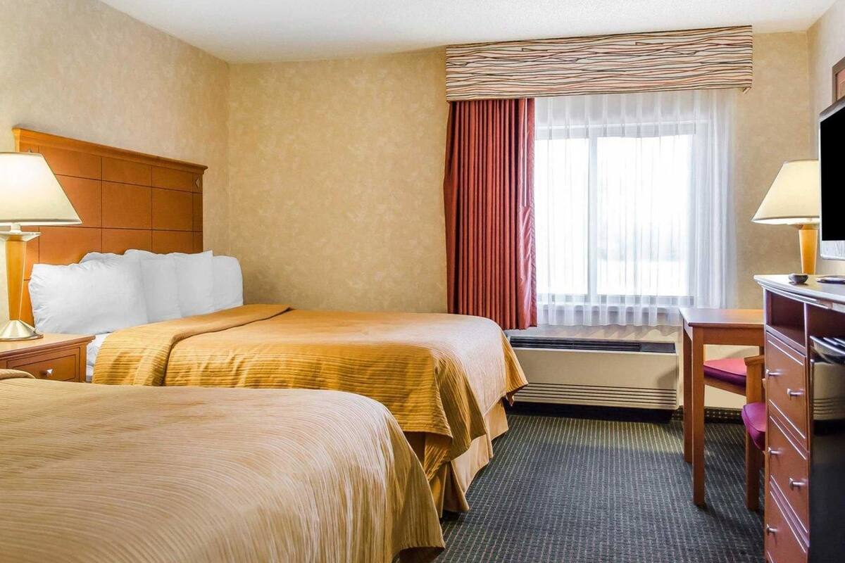 A hotel room with two beds, a desk, a dresser, and a large window with curtains.