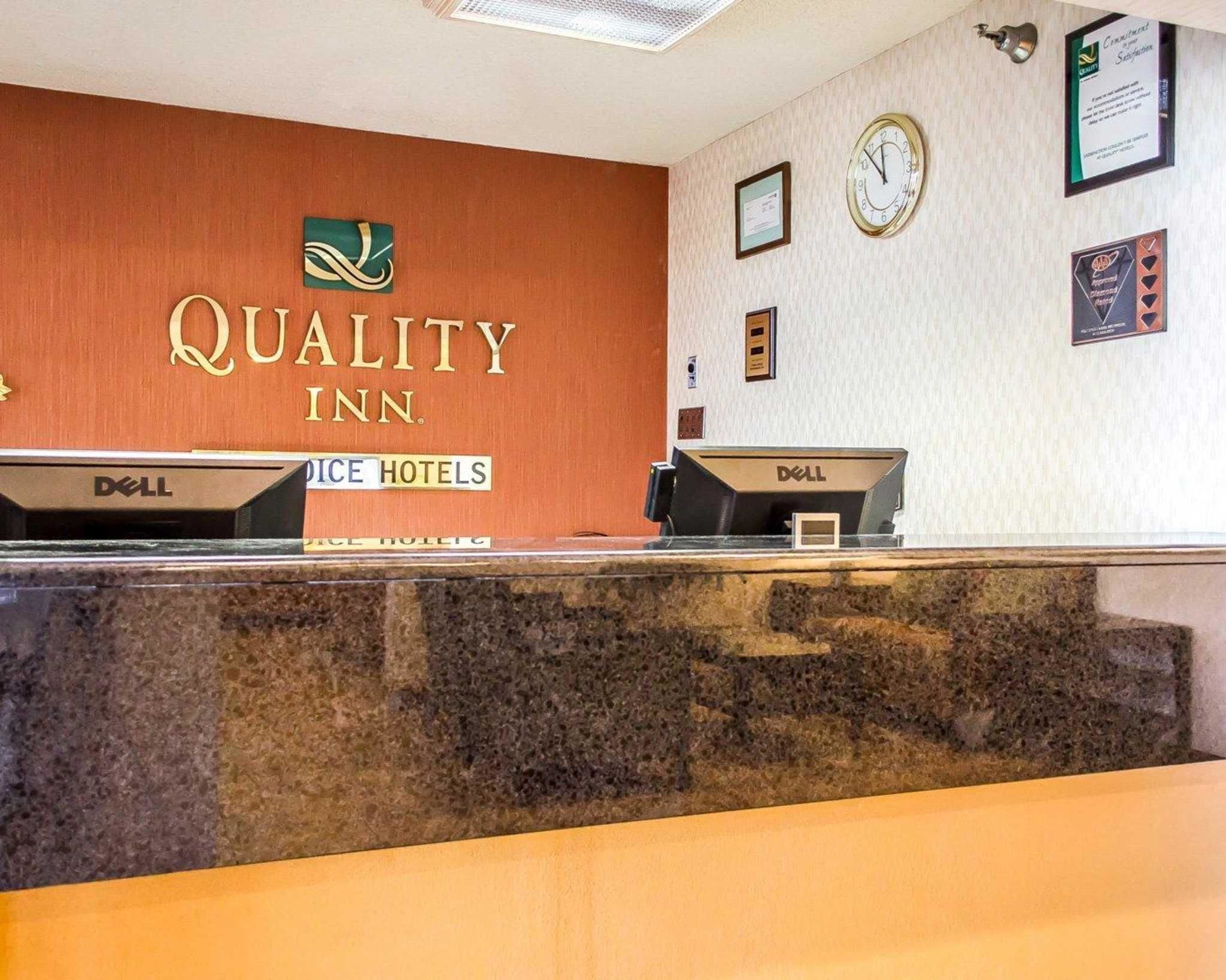 A hotel reception desk with a sign reading "Quality Inn." There are two computer monitors on the desk, and a clock on the wall.