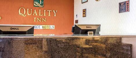 A hotel reception desk with a sign reading "Quality Inn." There are two computer monitors on the desk, and a clock on the wall.