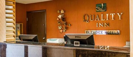 A hotel reception desk with a wooden finish, a sign reading "QUALITY INN BY CHOICE HOTELS," and a Dell computer monitor.