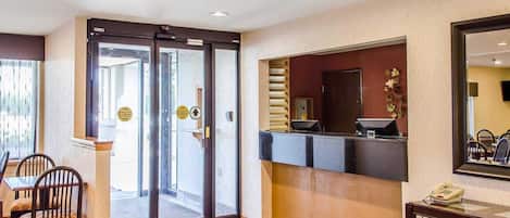 A hotel lobby with a reception desk, a seating area with chairs, a large mirror, and a door leading outside.