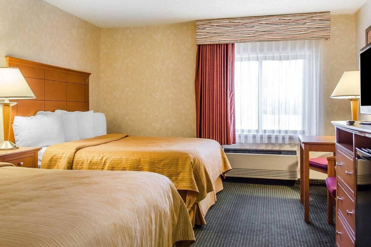 A hotel room with two beds, a desk, a dresser, a window with curtains, and a thermostat.