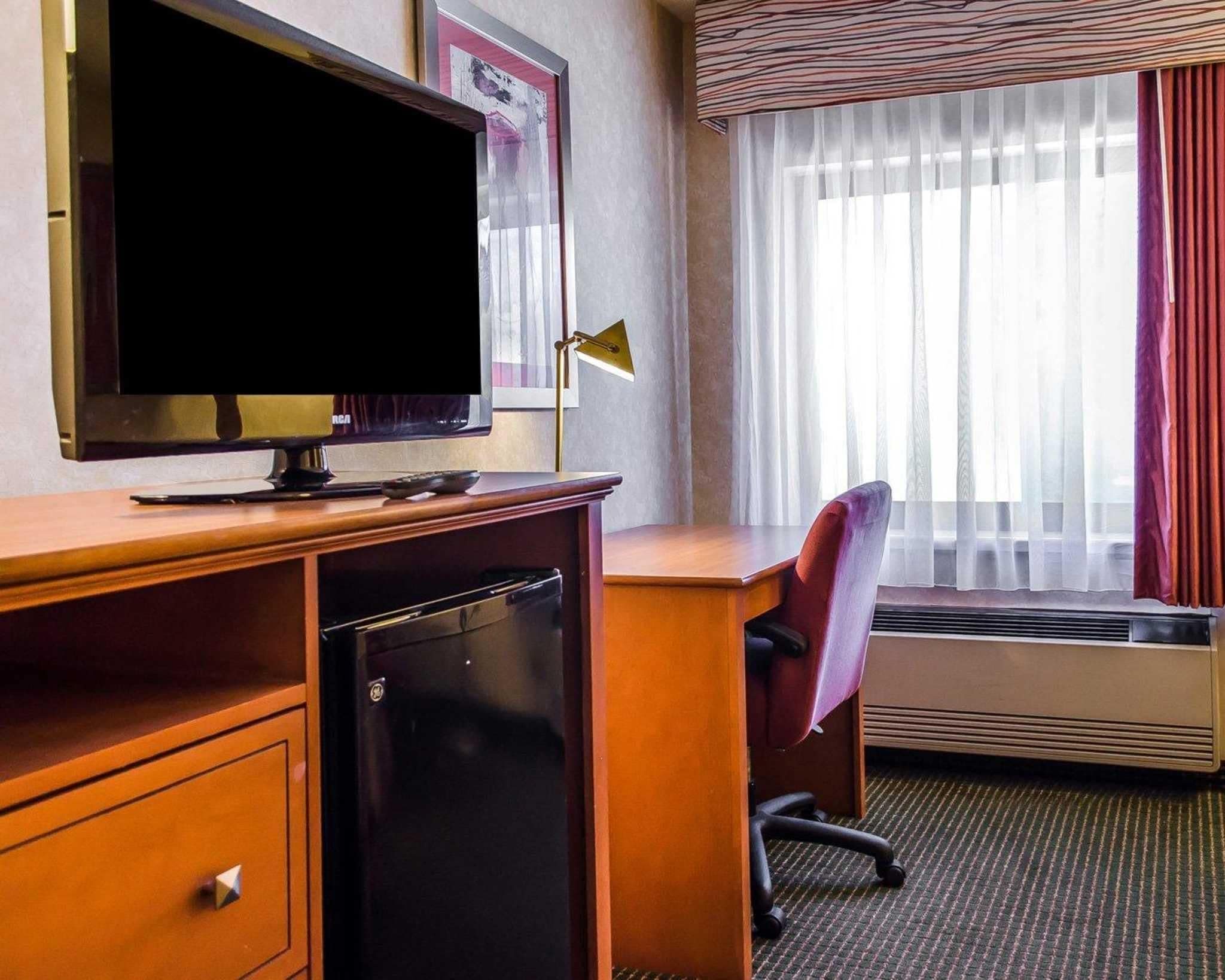 Et hotelværelse med et fladskærms-tv, et skrivebord med stol, et lille køleskab, et vindue med let gardin og en aircondition.