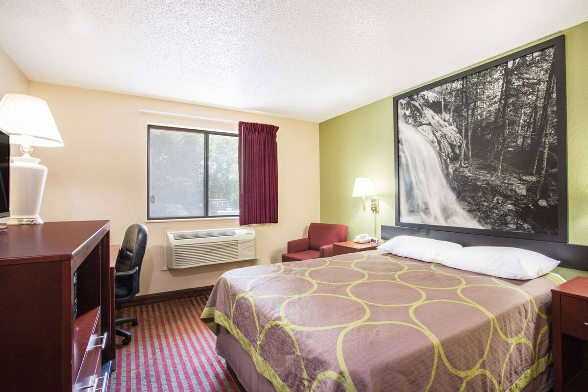 Une chambre d’hôtel comprenant un lit, un bureau, une chaise, un appareil de climatisation, une fenêtre et un grand tableau encadré au mur.