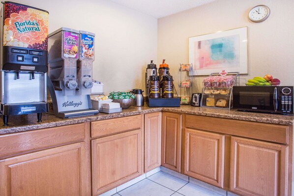 Una cocina con cafetera, dispensador de frutas, microondas y un reloj en la pared.