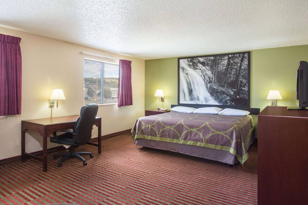 Une chambre d’hôtel avec un lit, un bureau, une chaise, deux lampes, une grande fenêtre avec des rideaux et un tableau représentant une cascade au mur.