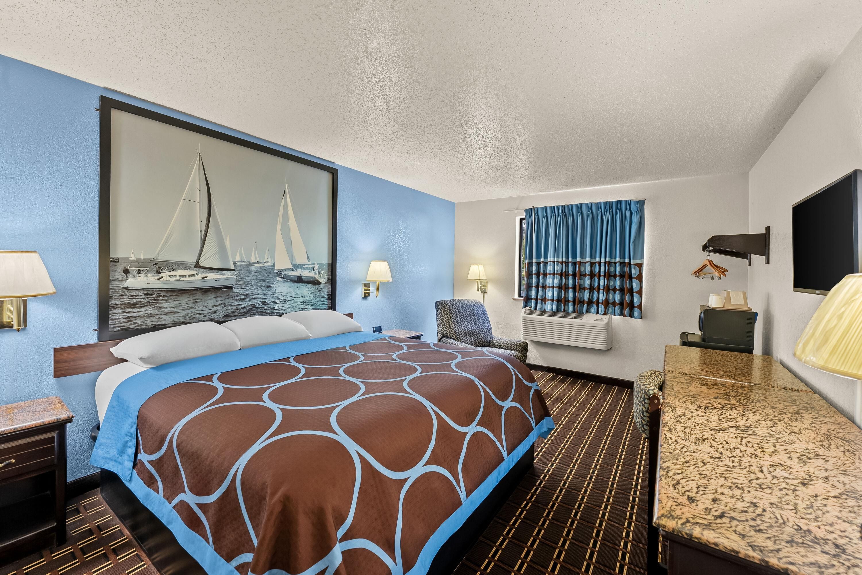 A hotel room with a large bed, a painting of sailboats, a desk with a chair, and a window with curtains.