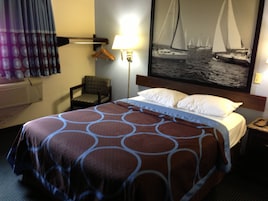 Habitación de hotel con cama, una silla, una lámpara, un cuadro de veleros y una ventana con cortinas.