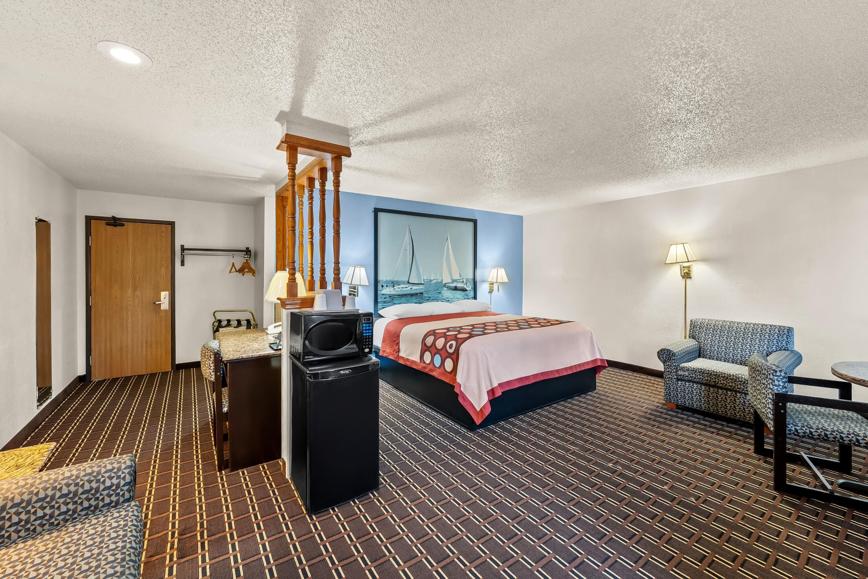 A hotel room with a bed, a desk, a chair, a microwave, and a lamp.