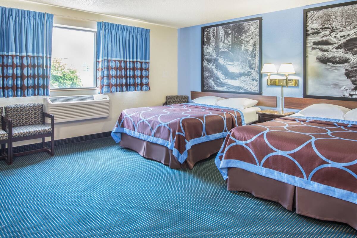 Une chambre d’hôtel avec deux lits, une fenêtre avec des rideaux, un climatiseur fixé au mur, une tête de lit avec une lampe et un grand tableau encadré au mur.