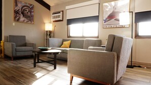 A living room with a sofa, armchair, and coffee table. There are two windows with blinds, a lamp, and a painting on the wall. - AmericInn by Wyndham Forest Lake (Forest Lake)