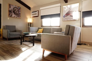 A living room with a sofa, armchair, and coffee table. There are two windows with blinds, a lamp, and a painting on the wall.