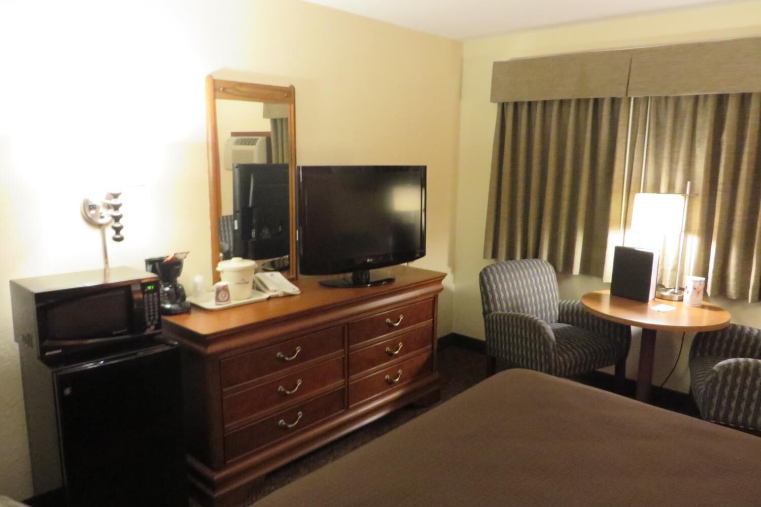 A hotel room with a bed, a TV, a desk with a lamp, a chair, a dresser, and a microwave.