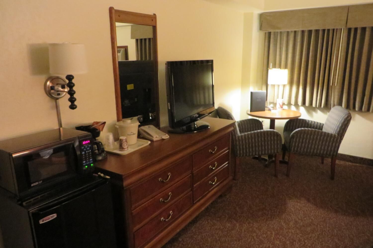 A hotel room with a microwave, a television, a coffee maker, a desk with a lamp, and two chairs.