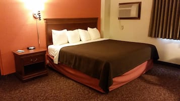 A hotel room with a bed, a nightstand, a lamp, an air conditioning unit, and a window with blinds.