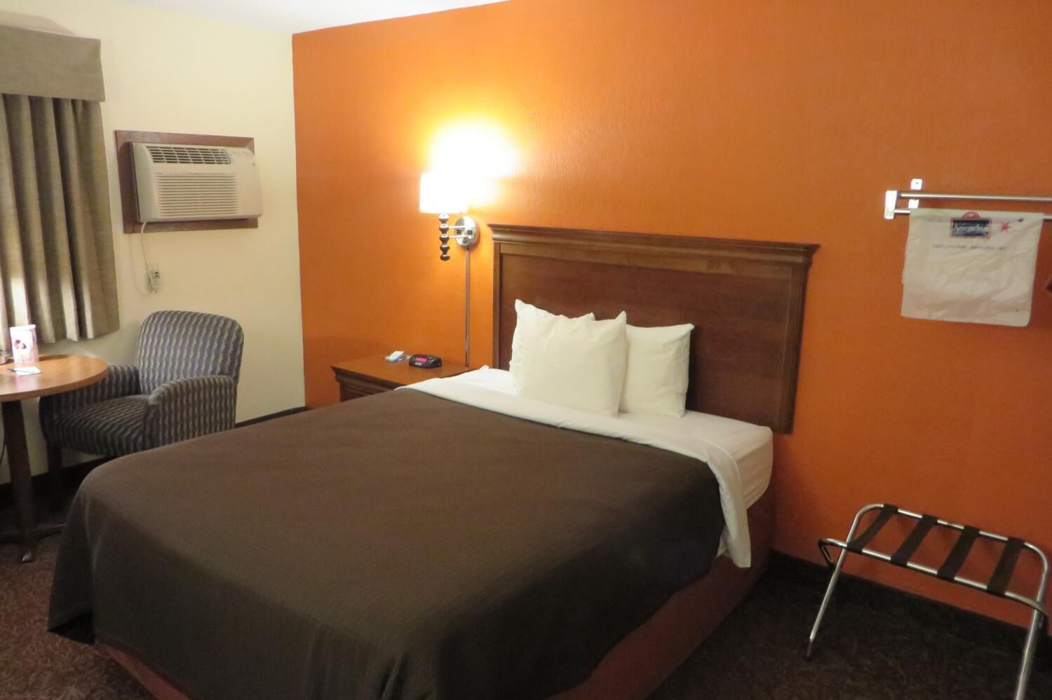 A hotel room with a large bed, a bedside table, a chair, an air conditioning unit, and a lamp.