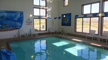 An indoor swimming pool with clear water, surrounded by windows and white chairs.