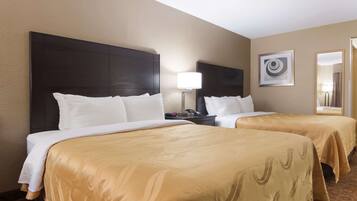 A hotel room with two beds, each with a dark headboard and white bedding. There is a bedside table with a lamp, a framed artwork on the wall, and a mirror.
