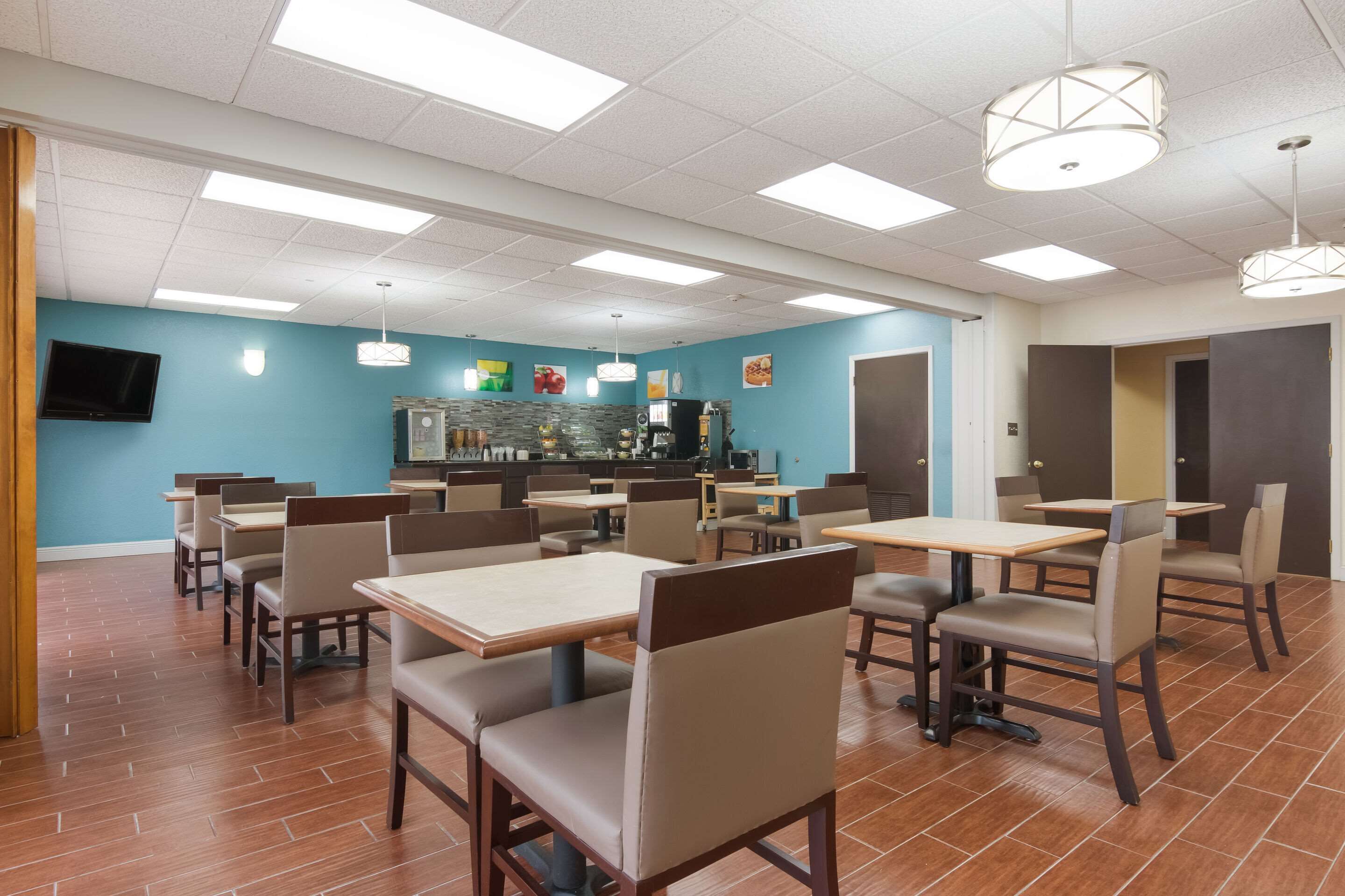 A dining area with tables and chairs, a kitchen in the background, and a television mounted on the wall.