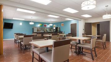 A dining area with tables and chairs, a kitchen in the background, and a television mounted on the wall.